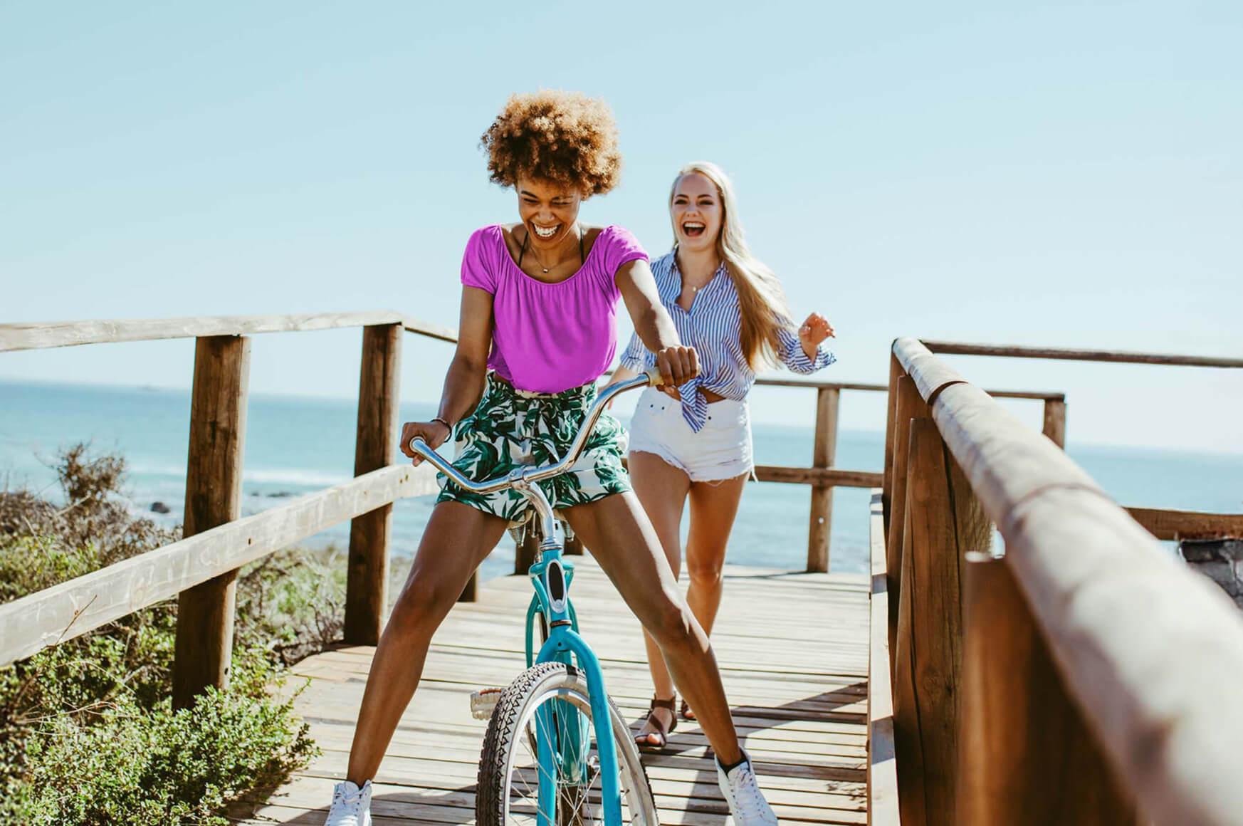 A woman rides a bike on a beach walkway as her friend laughs behind her