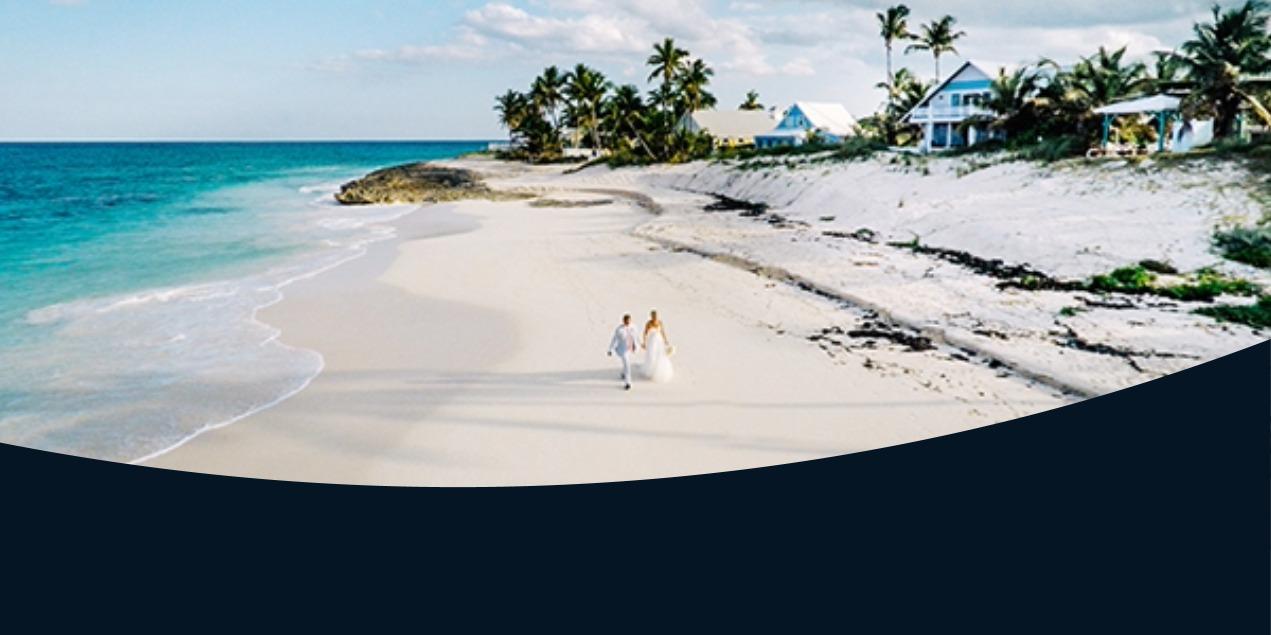 A bride and groom walk hand-in-hand down a wide, white-sand beach with turquoise water and palm trees in the background.