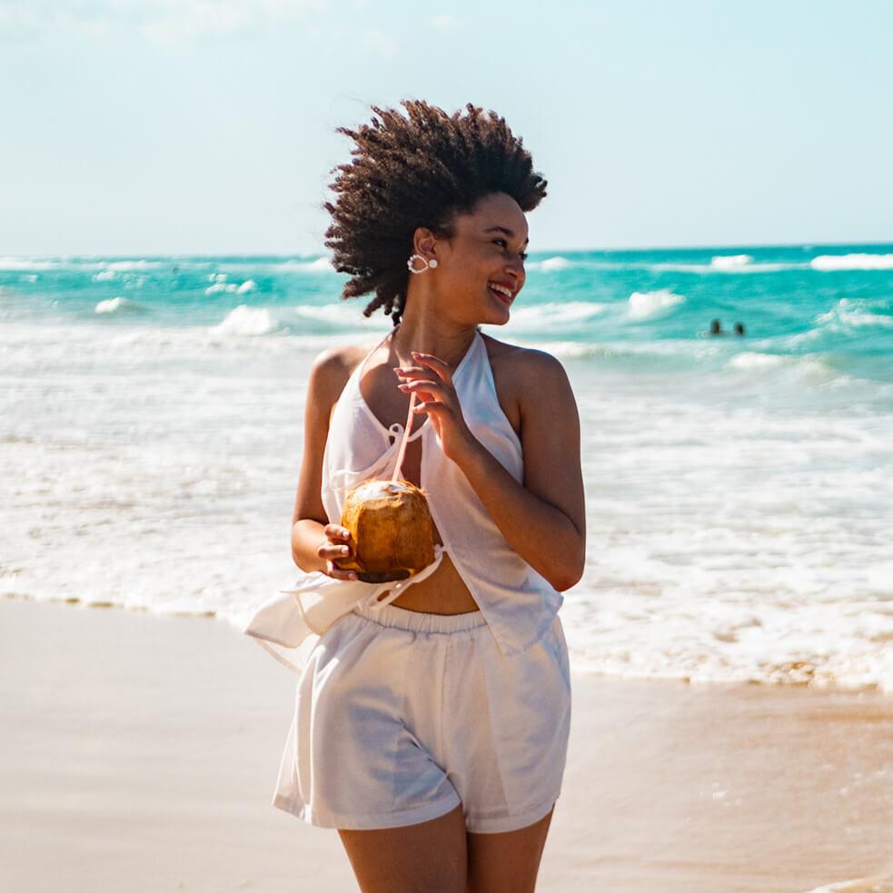 A women enjoys a drink from a coconut while walking along a beach with the ocean behind her